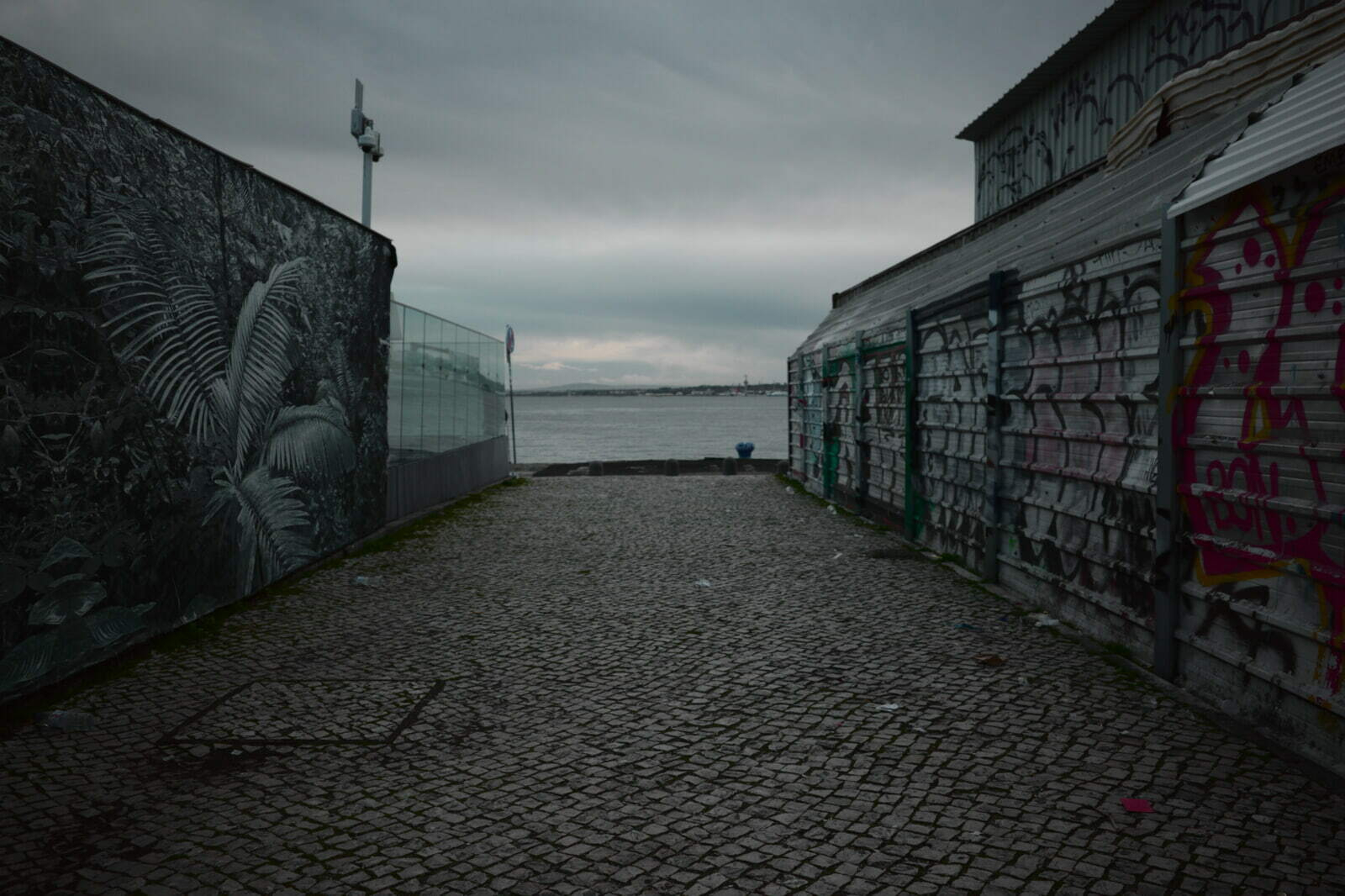 A cobblestone pathway flanked by graffiti-covered buildings leads to a waterfront view under a cloudy sky.