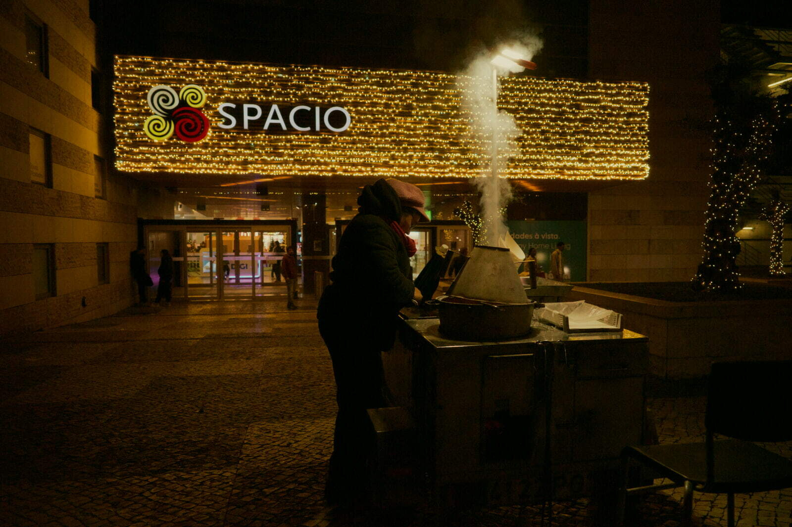 A street vendor stands by a roasting cart with steam rising, against a backdrop of a building adorned with illuminated decorations and the word SPACIO.
