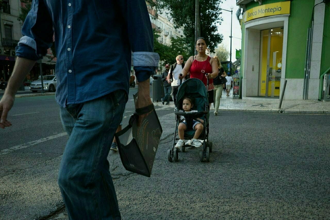 A woman pushing a child in a stroller walks on a city sidewalk near a bank named Banco Montepio.