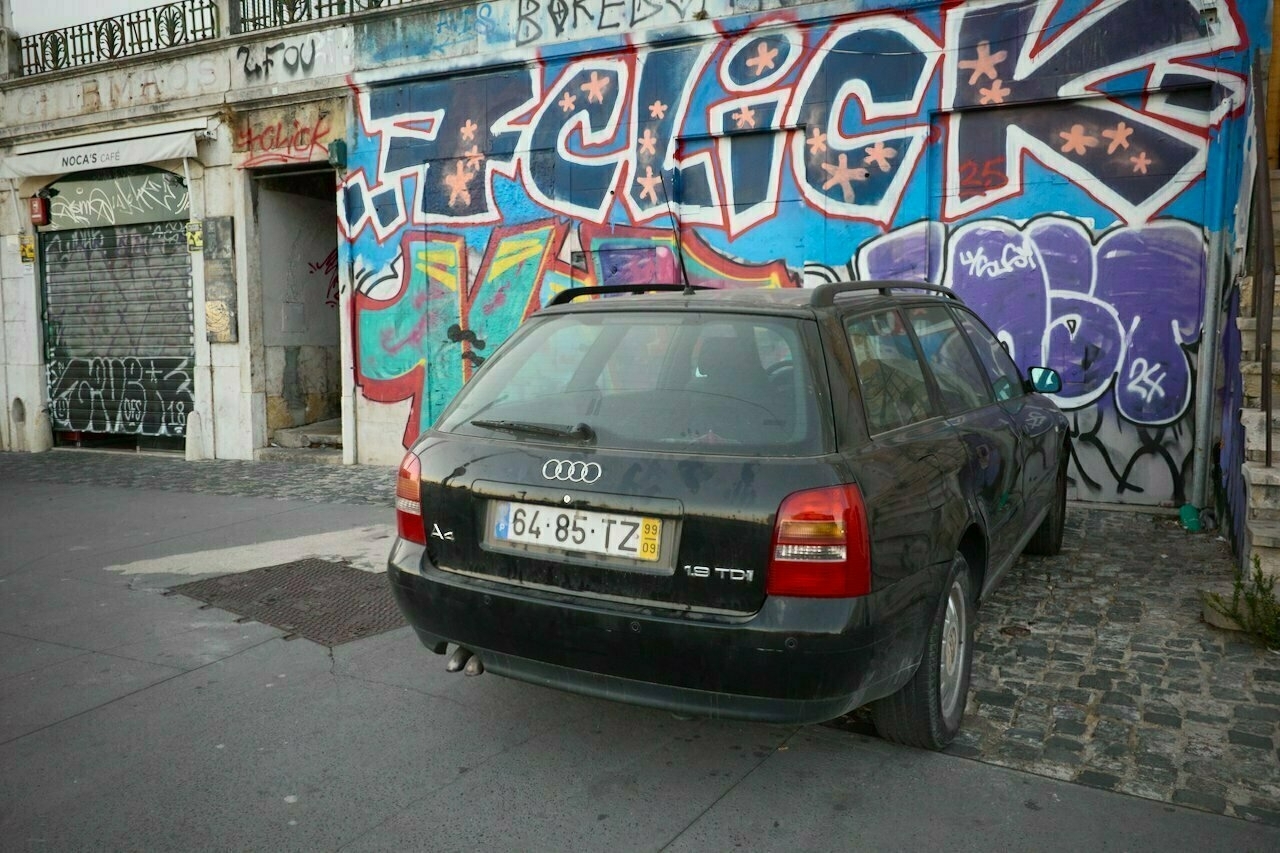 A black Audi station wagon is parked on a cobblestone street in front of a building covered in colorful graffiti.