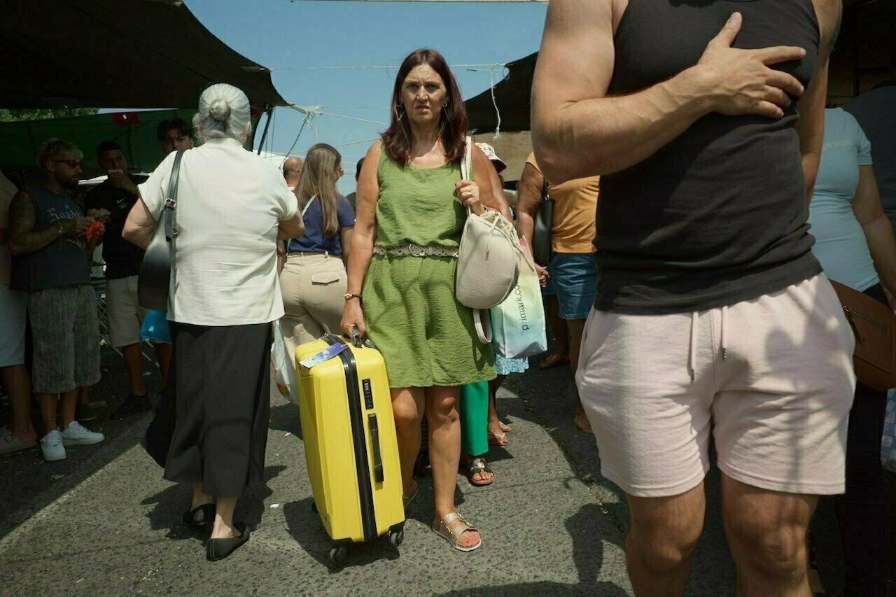 A woman in a green dress pulls a yellow suitcase through a crowded market or street area.
