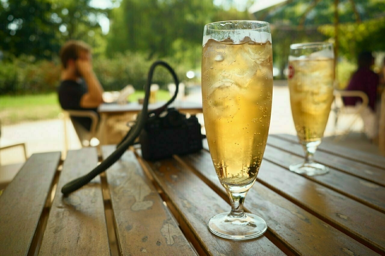 Two glasses of cider and a camera sit on a wooden outdoor table with people in the background.