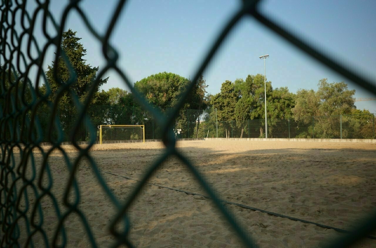 A sandy outdoor soccer field with a goalpost is viewed through a chain-link fence, surrounded by trees under a clear sky.