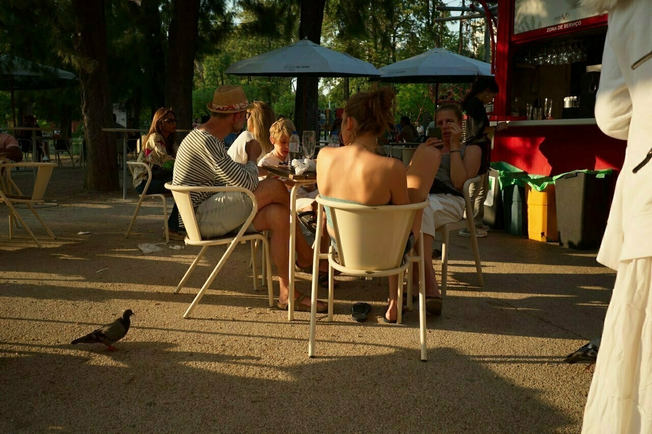 People are sitting at outdoor tables under umbrellas, enjoying the sunny weather, with a few pigeons nearby.