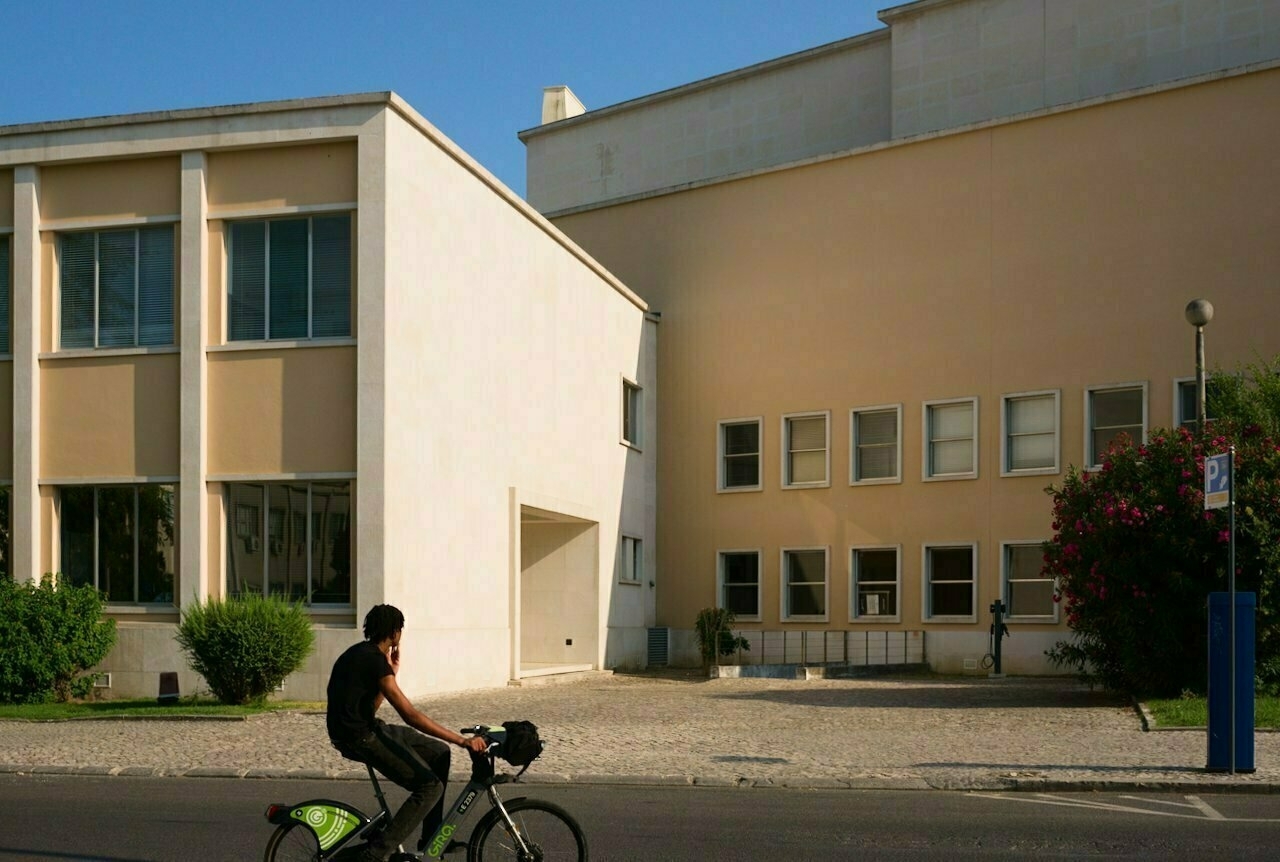 A person rides a bicycle past modern buildings with large windows and a small sidewalk.