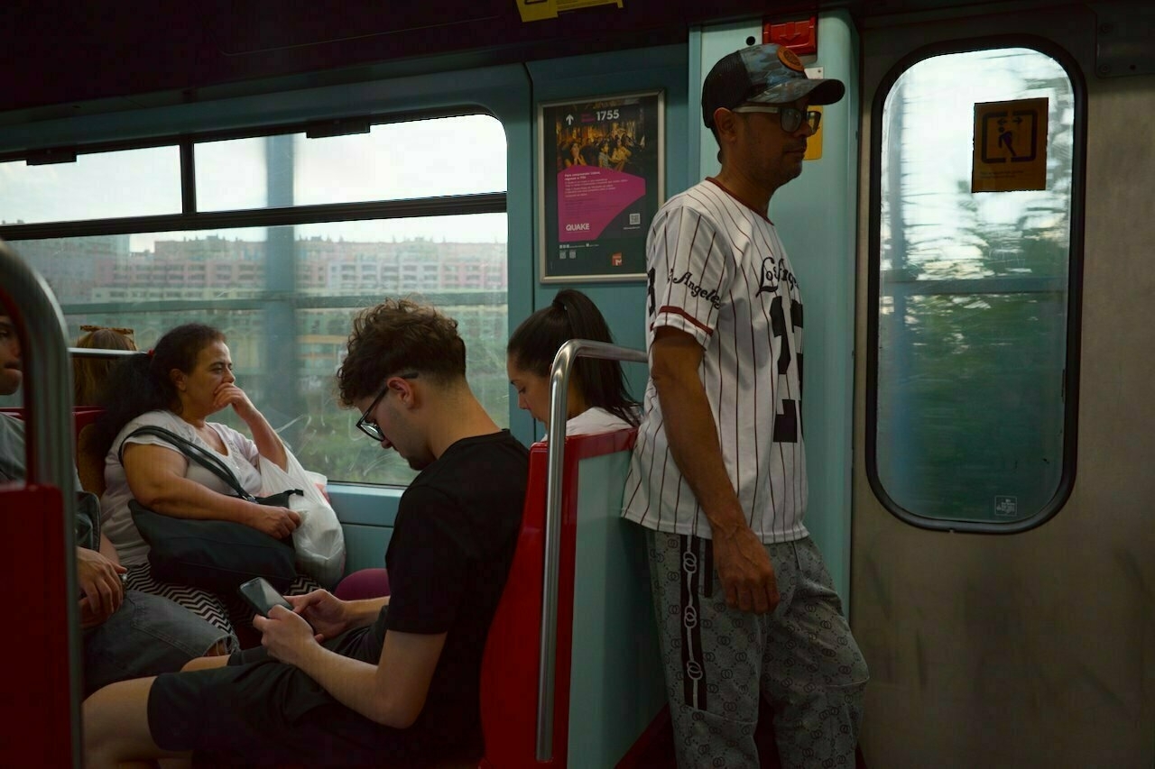 Passengers are seated and standing inside a moving train, with sunlight streaming through the windows.