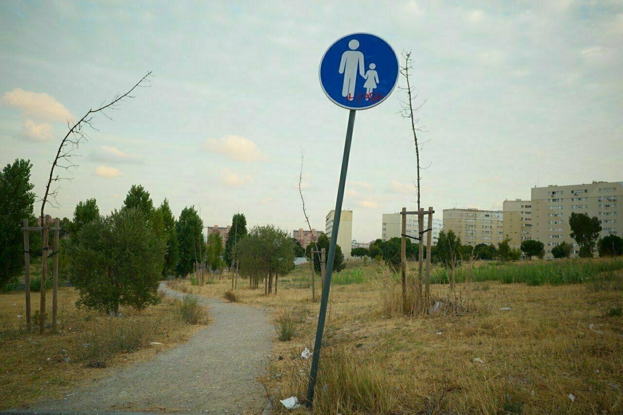 A footpath in a grassy area features a tilted pedestrian sign for adults with children, surrounded by sparse trees and distant apartment buildings.