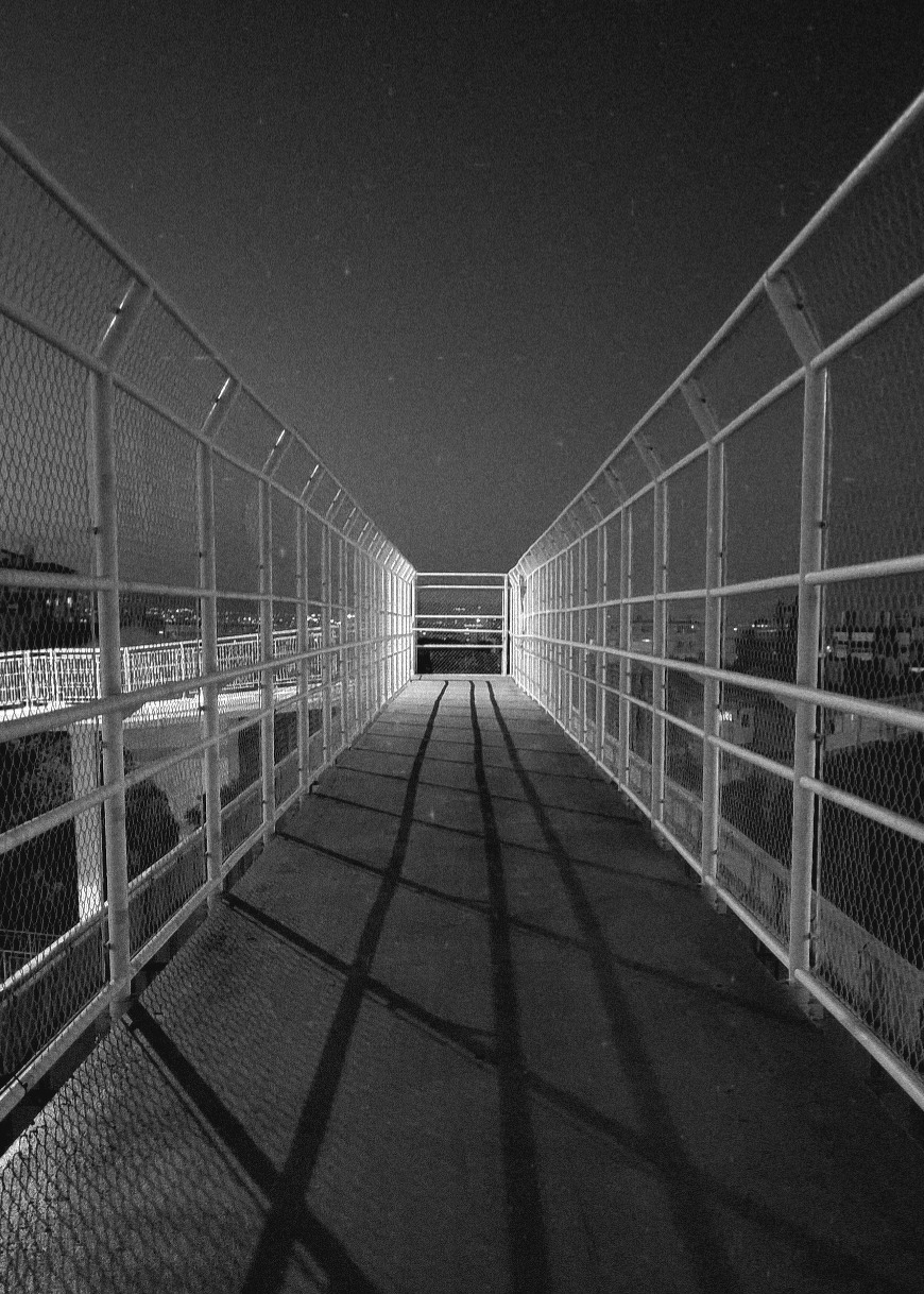 A long, illuminated pedestrian bridge with metal railings casts shadows on the ground at night.