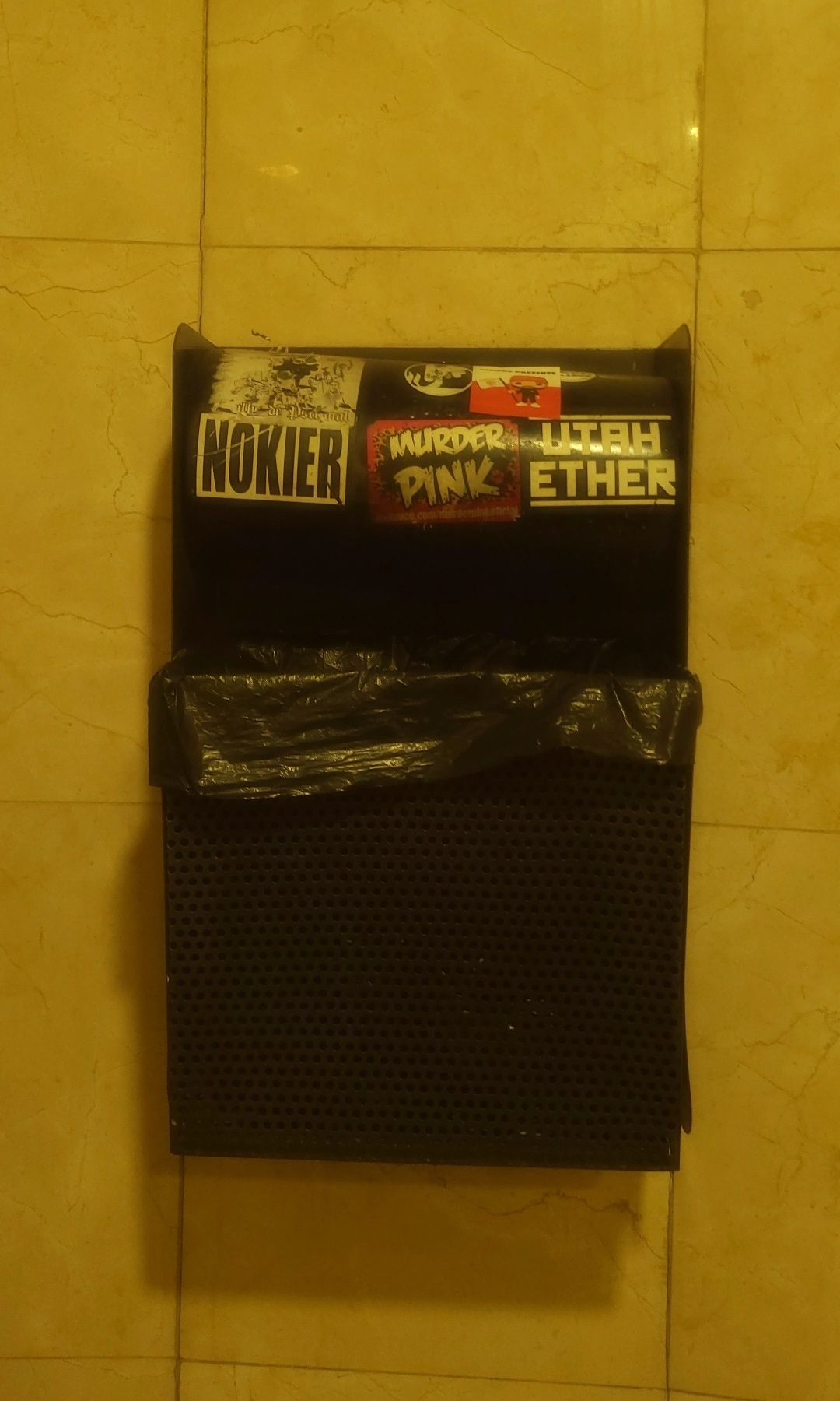 A wall-mounted black trash bin is covered with stickers and has a plastic bag inside, set against a beige tiled background.
