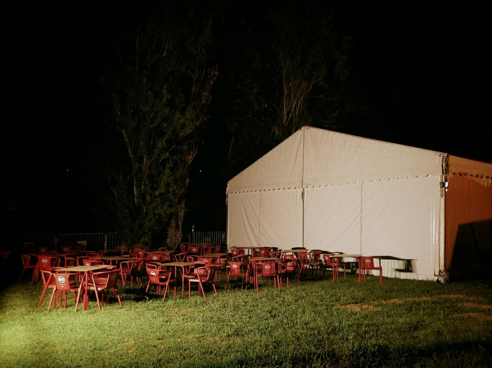 An outdoor seating area with red chairs and tables is illuminated at night next to a large white tent.