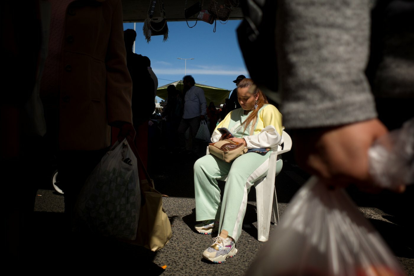 A person sitting on a chair outdoors is feeding a baby, surrounded by people carrying shopping bags.
