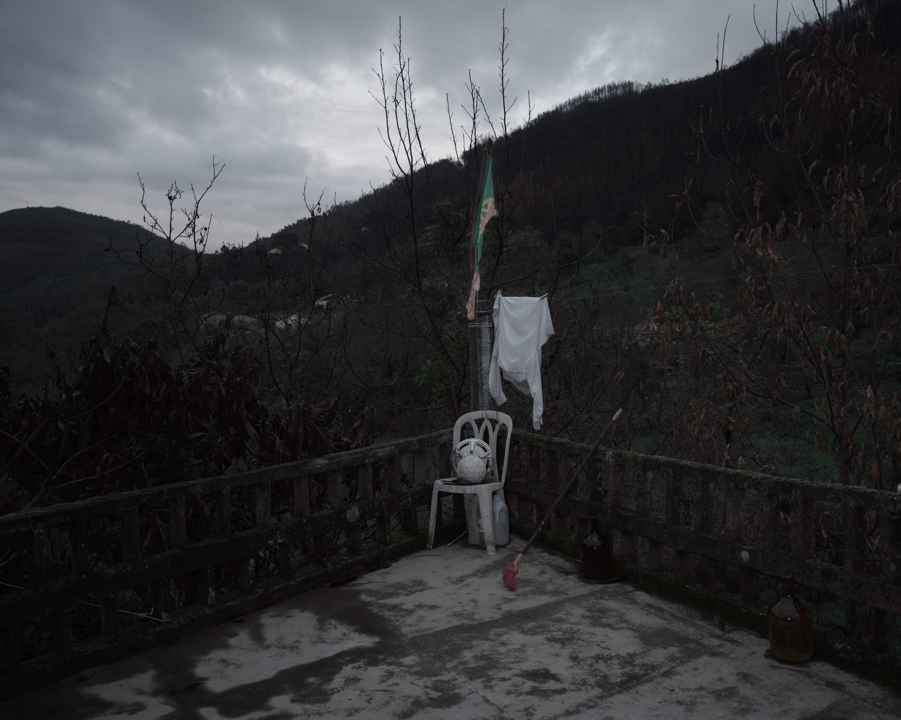 A moody, overcast scene depicts a concrete terrace with a white plastic chair, a broom, a hanging cloth, and a flag, set against a backdrop of mountains and leafless trees.