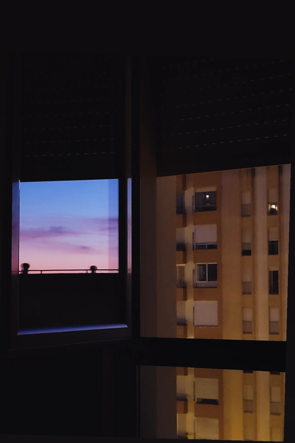 A view through a window shows a colorful sunset sky on one side and a beige apartment building on the other.