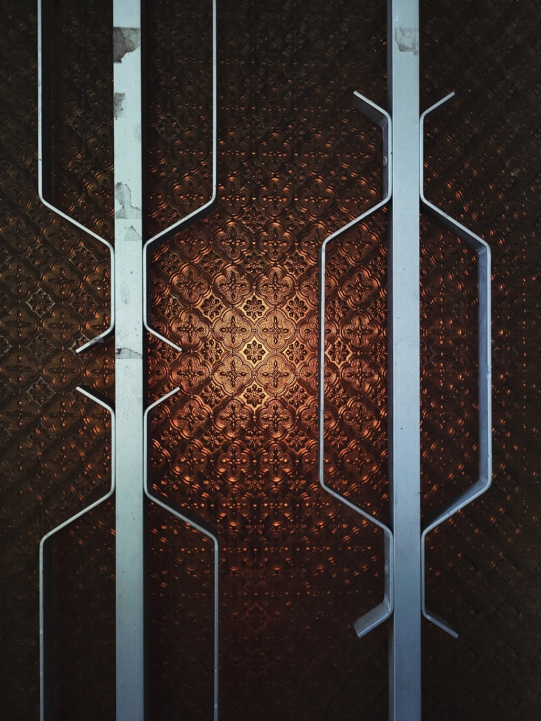 Vertical metal frames with irregular shapes are positioned against a patterned, embossed brown textured background, illuminated by a warm light source.