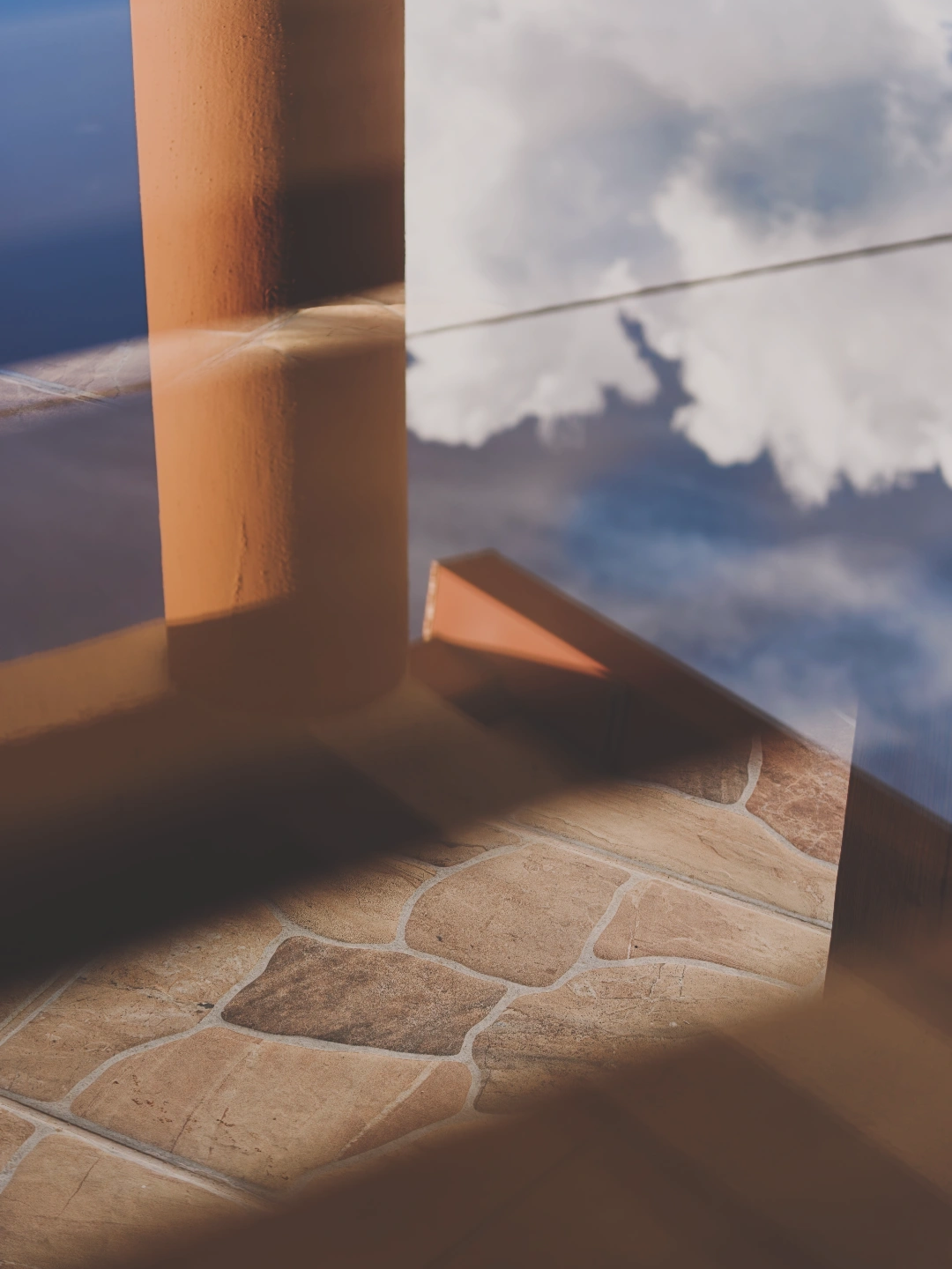 A double-exposure or reflective photograph blending a stone-tiled floor and an orange architectural column with a bright blue sky filled with white clouds. Dark, geometric shadows stretch across the multi-colored tan and brown flagstone flooring on the bottom half, while a thin, faint horizontal line cuts across the reflected sky in the upper portion.