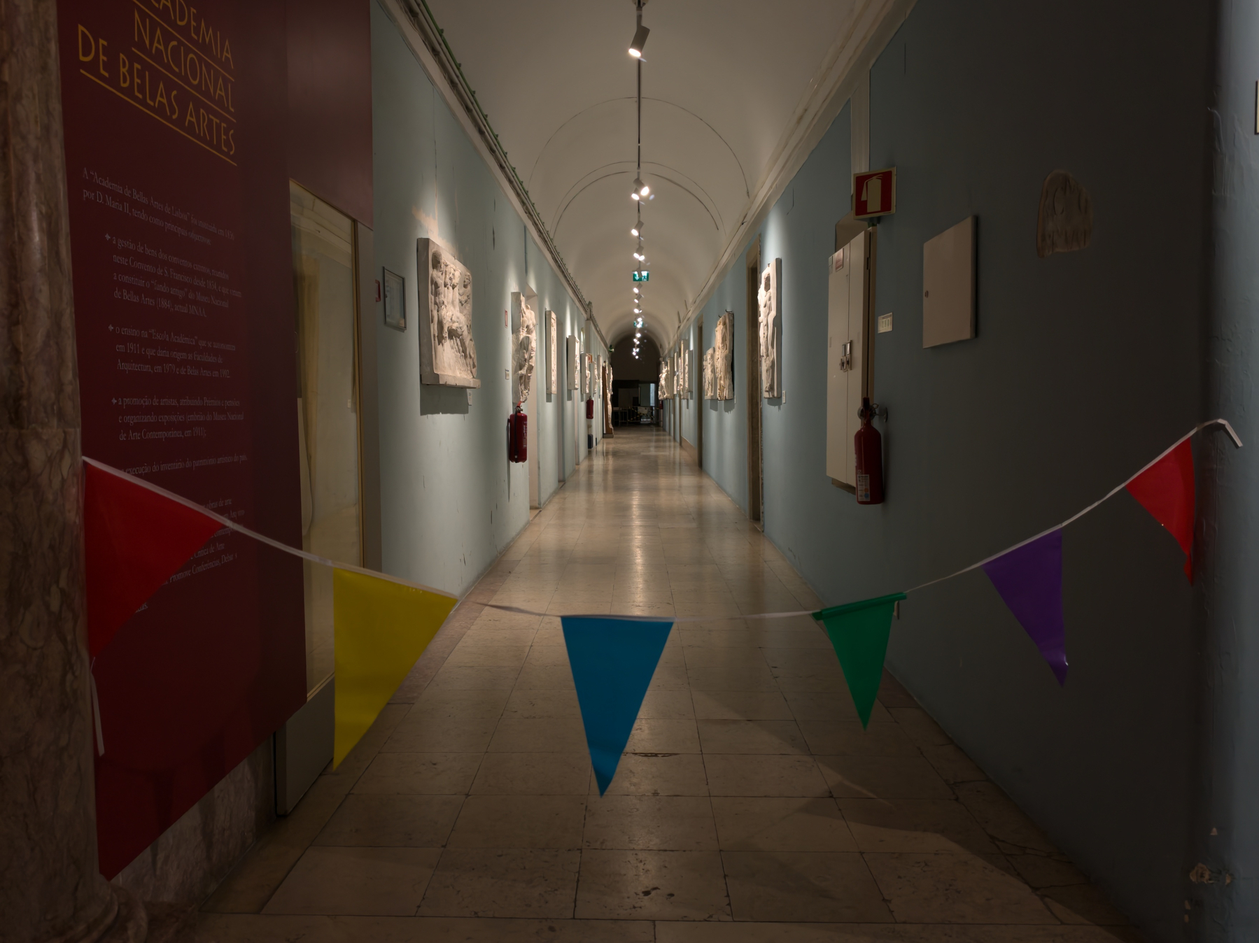 A long, perspective-view corridor inside an art academy with light blue walls and a white vaulted ceiling. High on the left wall, a dark red sign features the text “ACADEMIA NACIONAL DE BELAS ARTES”. Classical white plaster relief sculptures are mounted at intervals along both walls. A colorful string of triangular pennant bunting—with yellow, blue, green, and purple flags—hangs across the foreground. The hallway is illuminated by overhead track lighting, leading toward a darker doorway at the far end.
