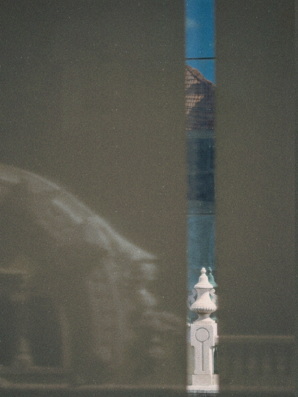 A narrow vertical reflection on a dark surface reveals part of a traditional rooftop and an ornamental stone post against a clear blue sky.