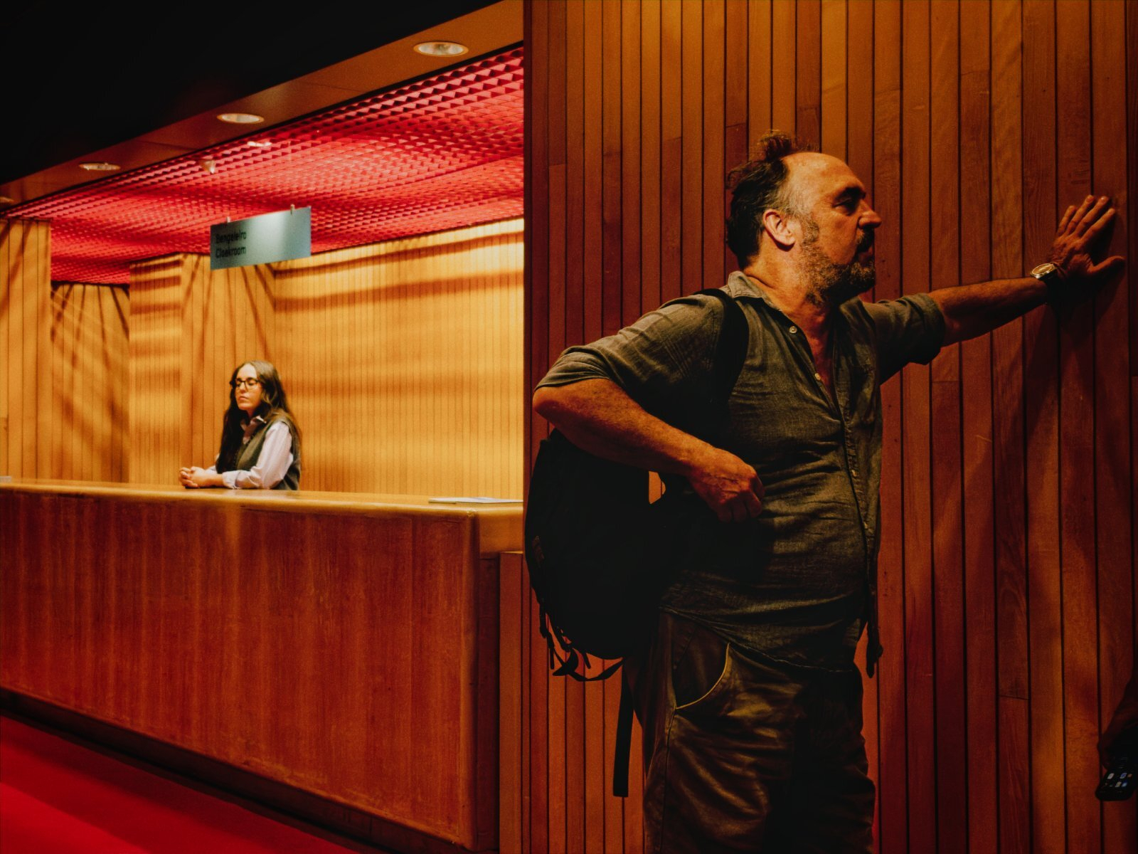 A man with a backpack leans against a wooden wall while a woman stands behind a counter in a dimly lit interior space.