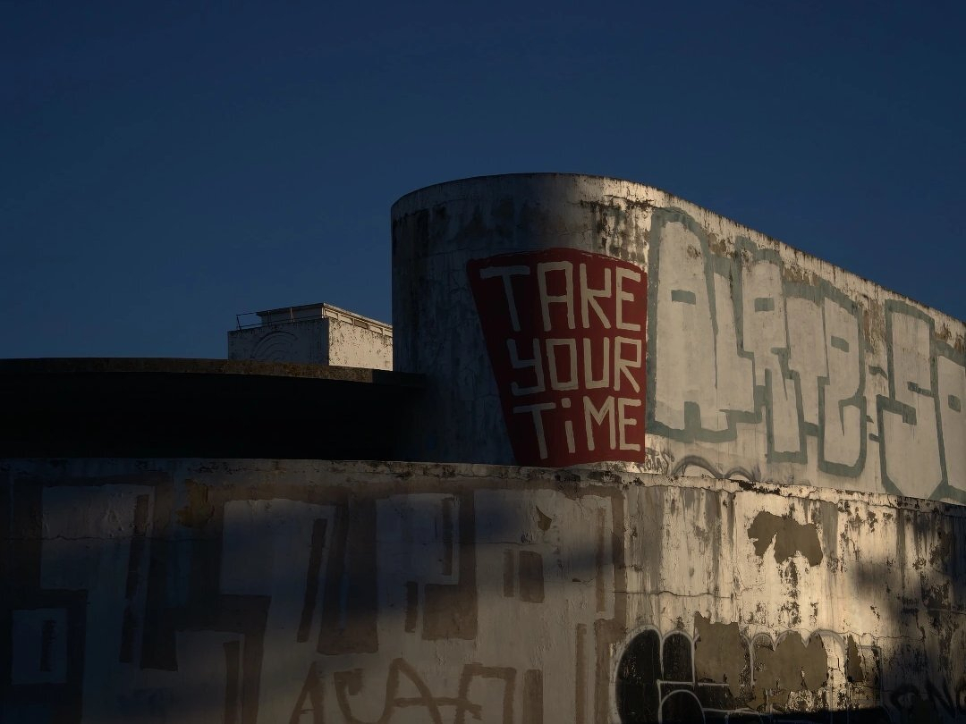 A weathered concrete building features graffiti, including a prominent red and white piece that says TAKE YOUR TIME.