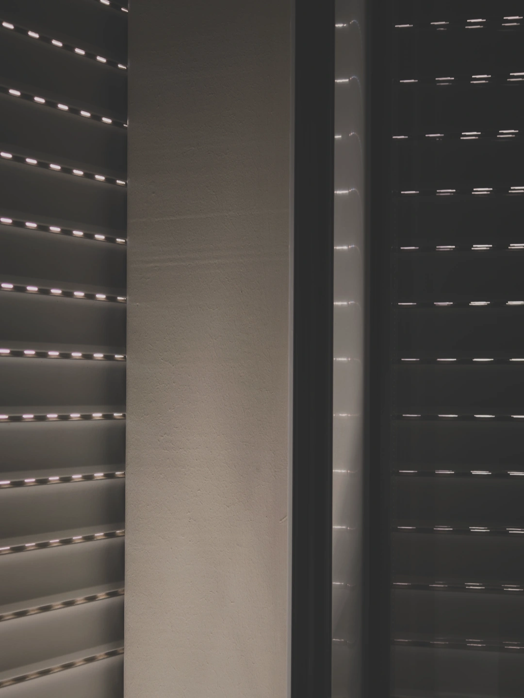 The image shows a window fitted with horizontal slat roller shutters or blinds that are partially closed, allowing thin, parallel lines of light to filter into a dimly lit interior space next to a plain wall.