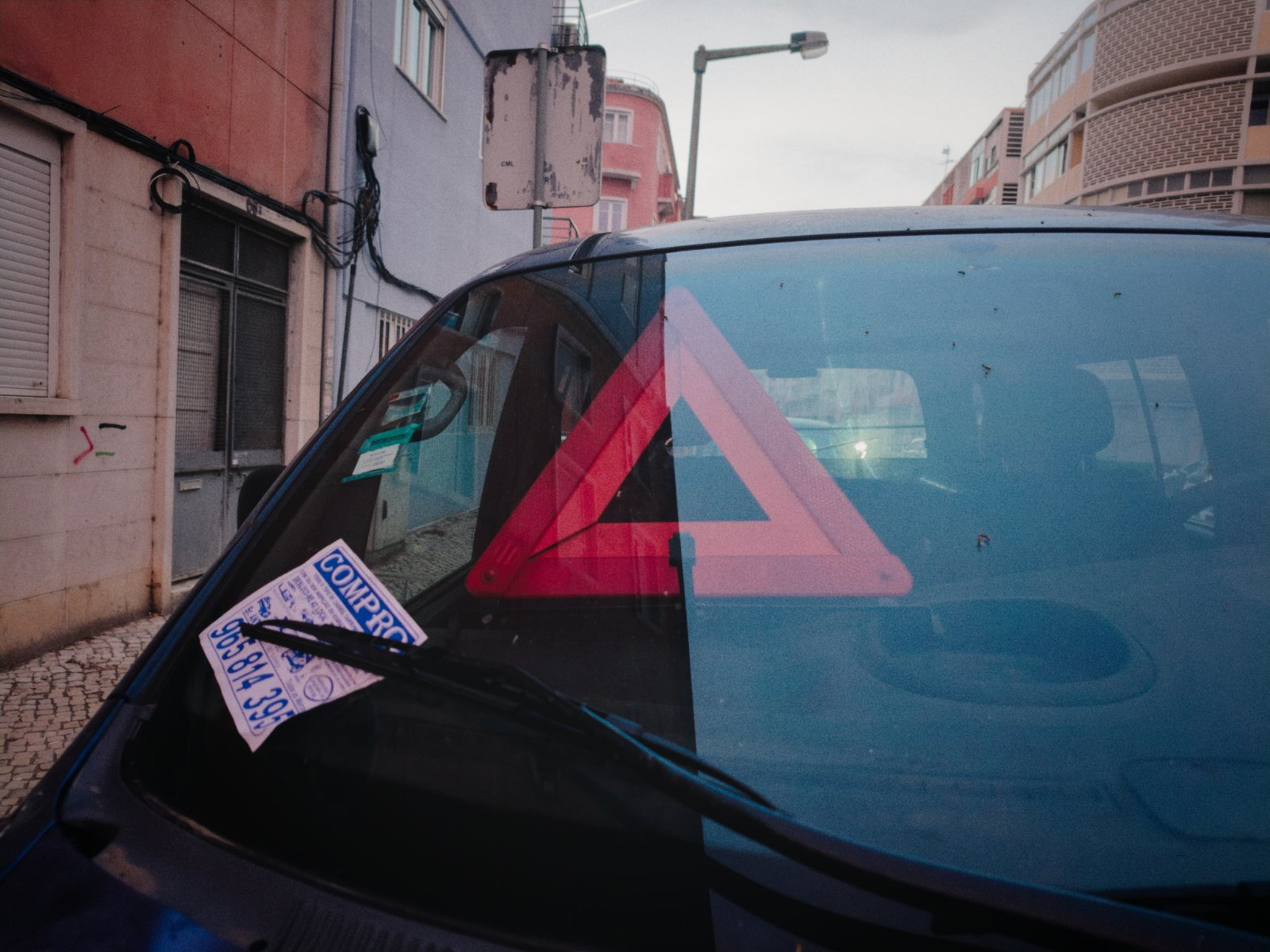 Windshield of a car parked on a city street, featuring a red emergency warning triangle placed on the dashboard and a flyer stuck under the windshield wiper.