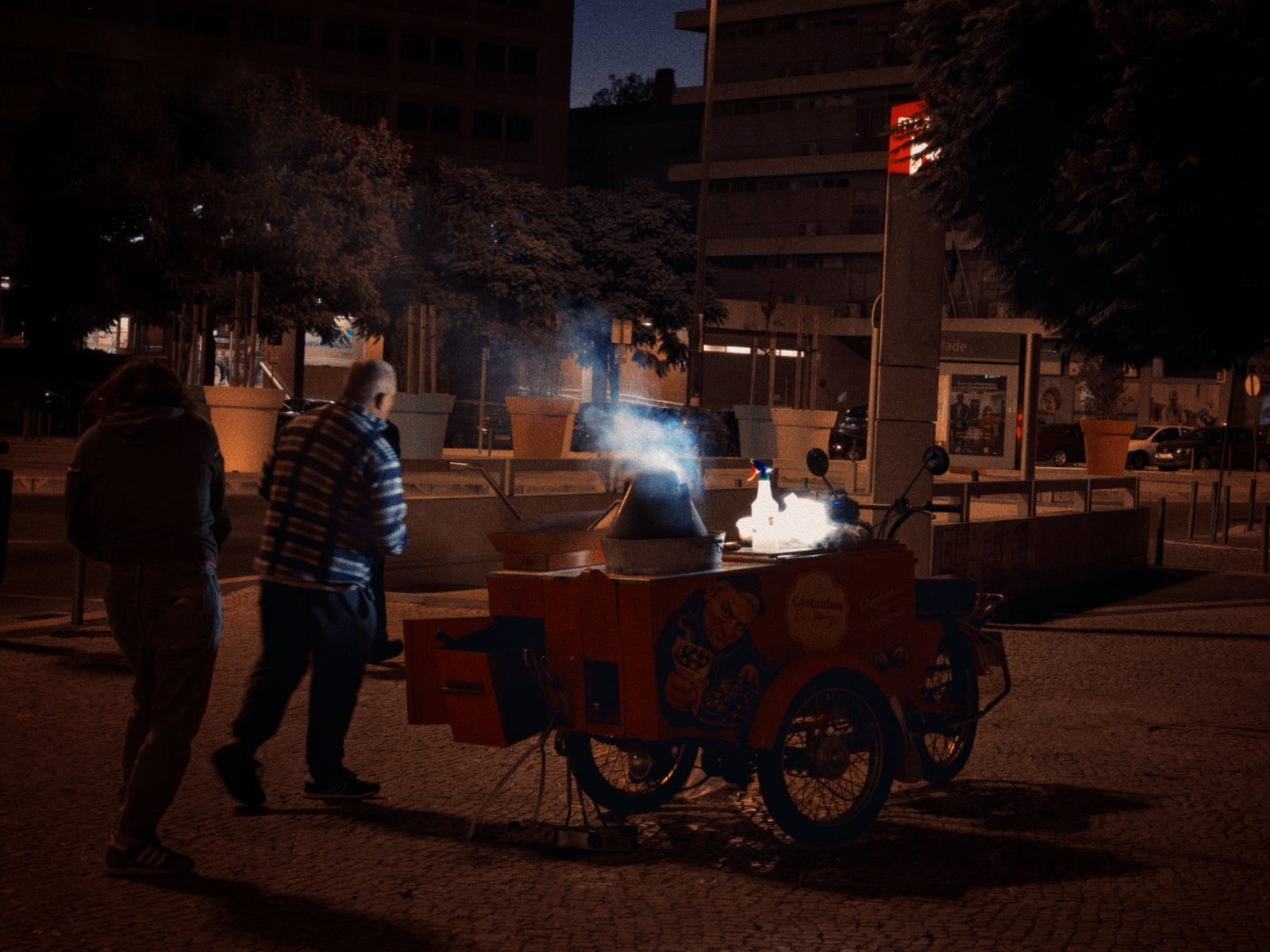Two people are walking past a street vendor's cart emitting smoke during nighttime in an urban setting.