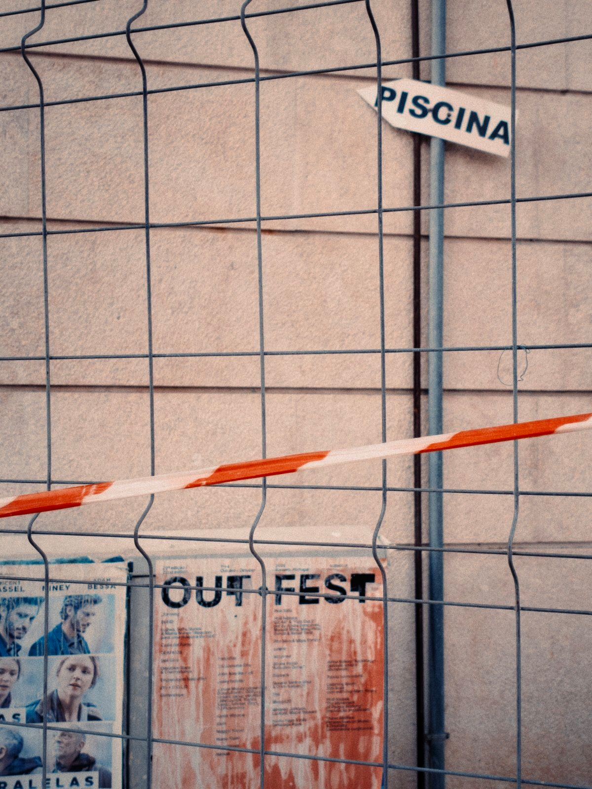 A metal fence with a red and white tape blocks the view of a wall featuring a poster with the title OUT FEST and a partially visible sign pointing towards a PISCINA.