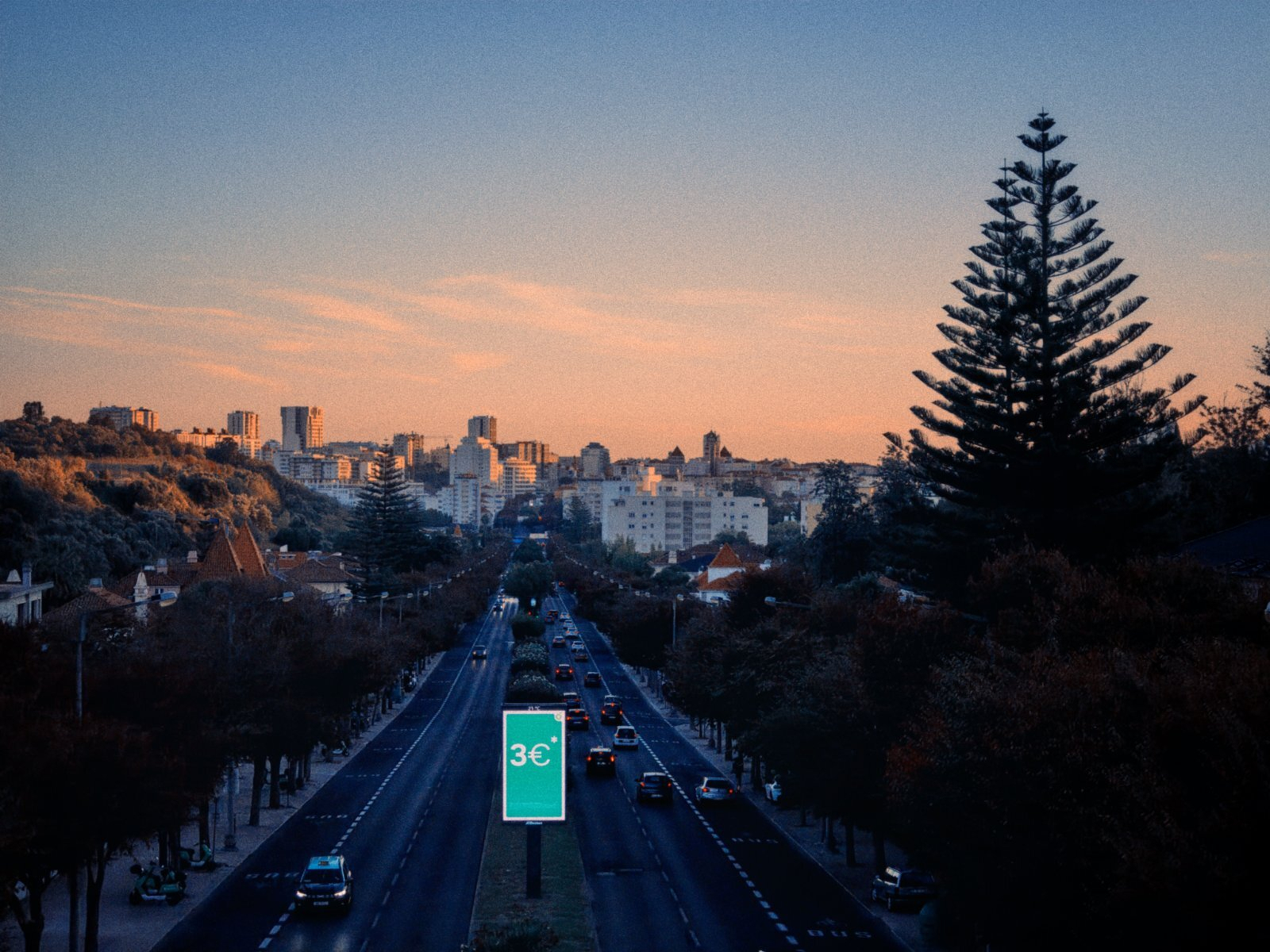 A cityscape is shown during sunset with a busy road lined by trees leading towards a cluster of buildings in the distance.