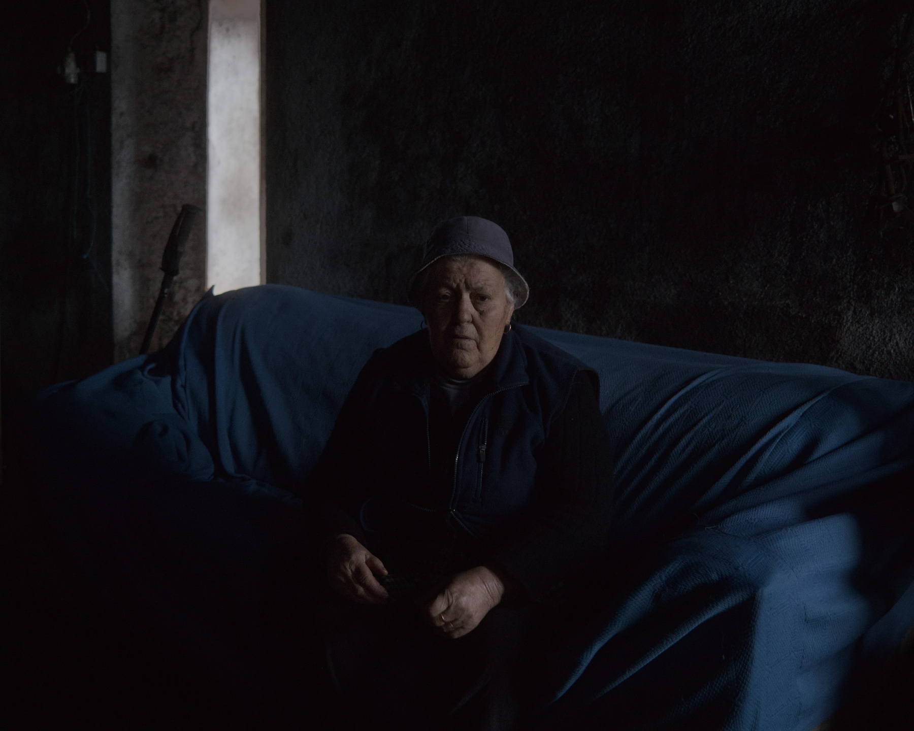 An elderly person wearing a hat sits on a couch draped with a blue cover, in a dimly lit room.