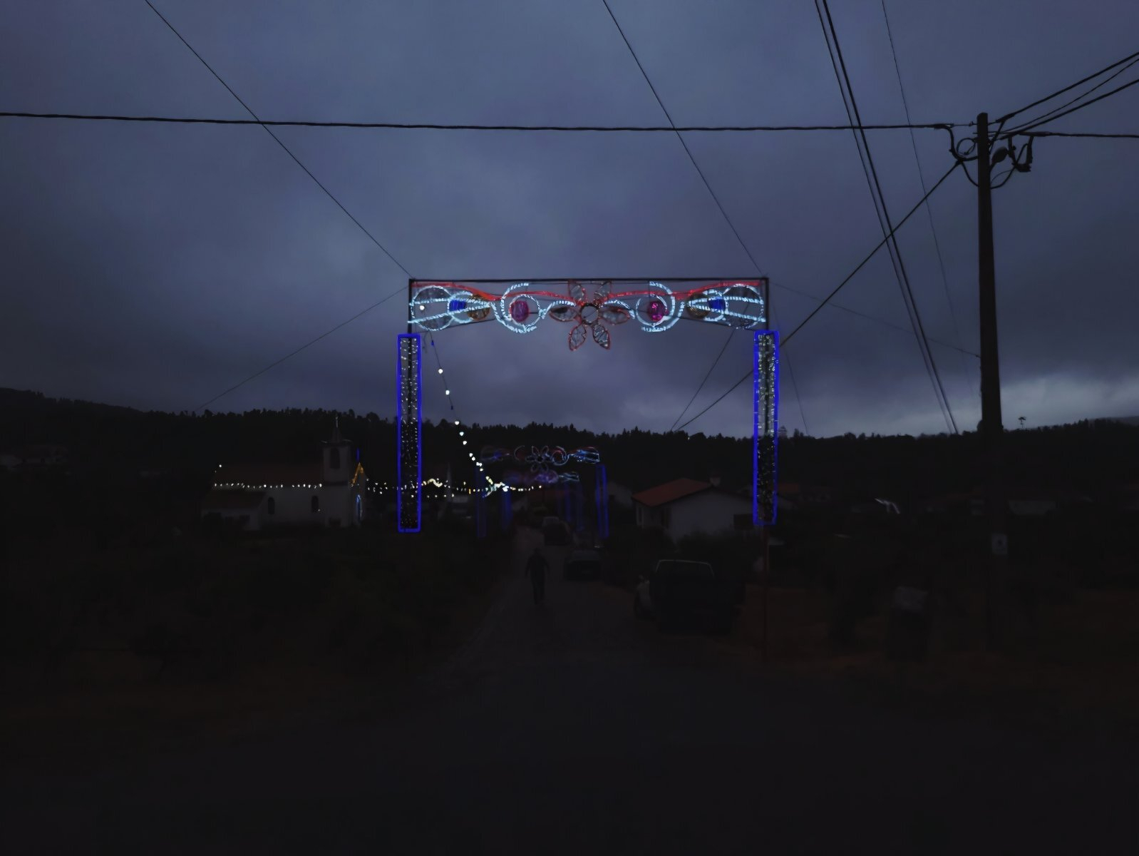 Decorative lights are strung across a dark, cloudy sky over a rural road, with faint outlines of buildings and trees in the background.