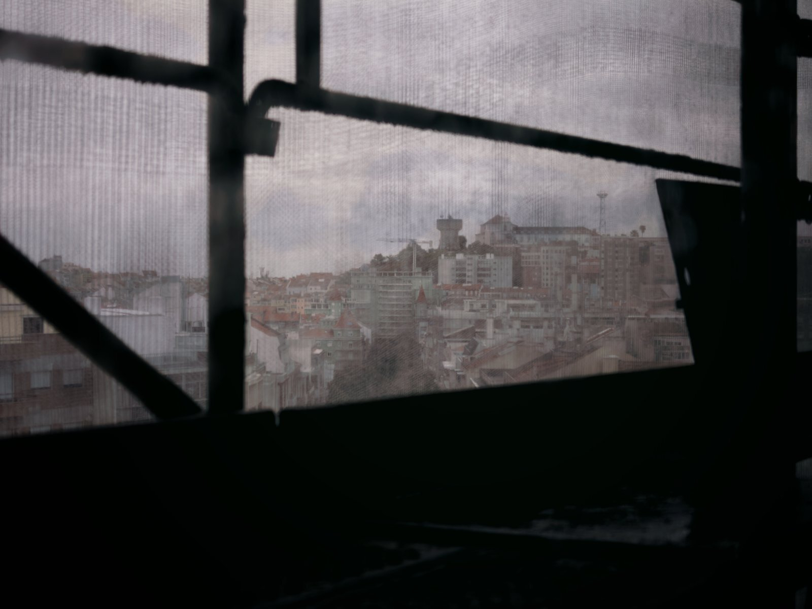 a view of the city of Lisbon, Portugal, framed through a window with a mesh screen, looking toward the historic upper districts and church towers