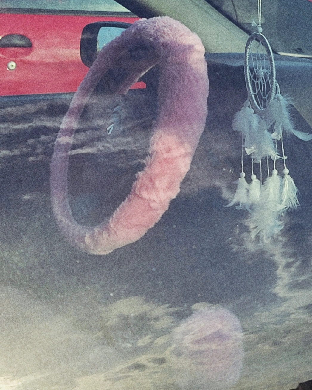 A car's steering wheel is covered with a pink fluffy cover, and a dreamcatcher hangs from the rearview mirror, with a reflection of clouds on the window.