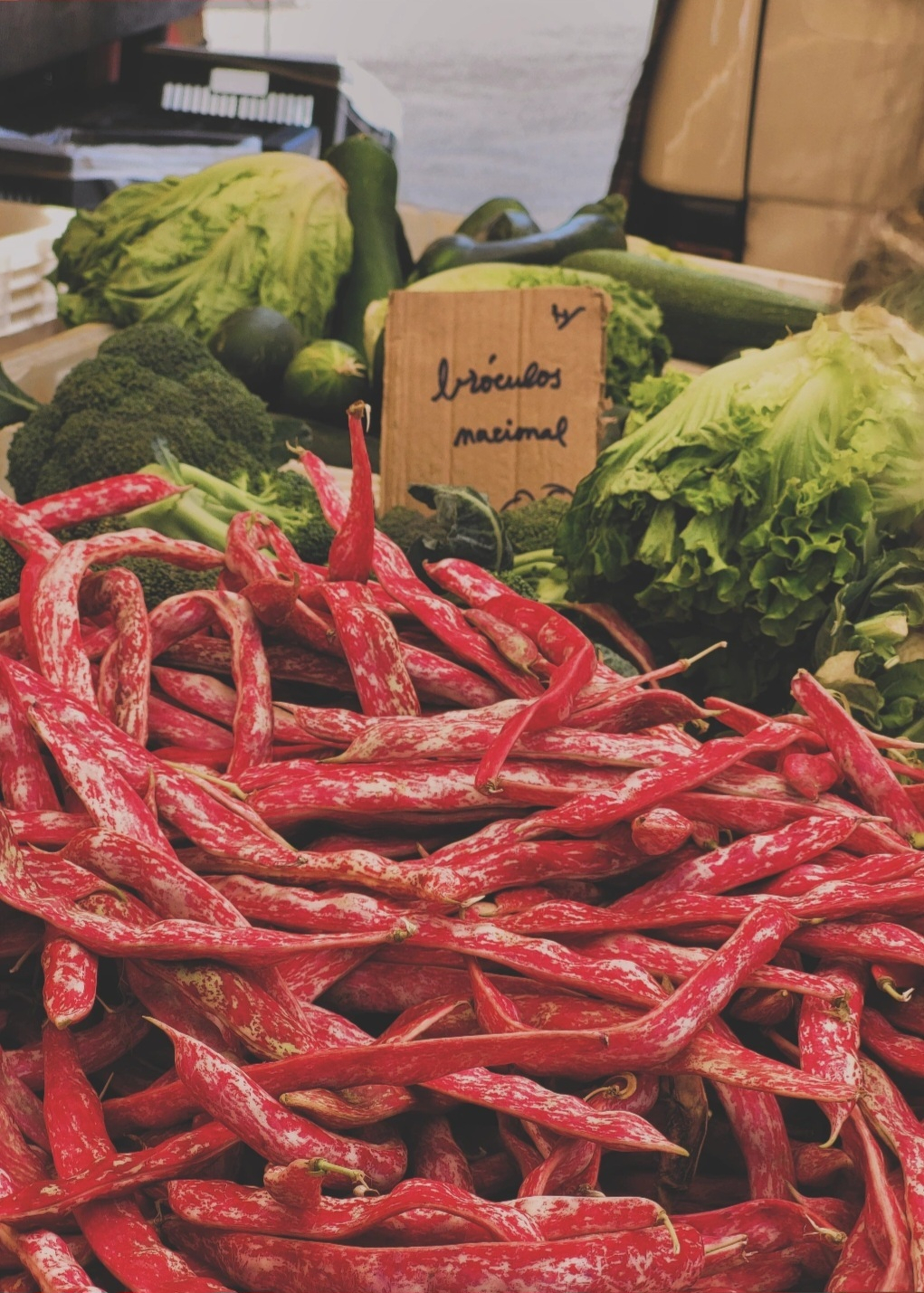 A market display features a large pile of red-speckled beans in the foreground, with leafy greens, broccoli, and a handwritten sign in the background.