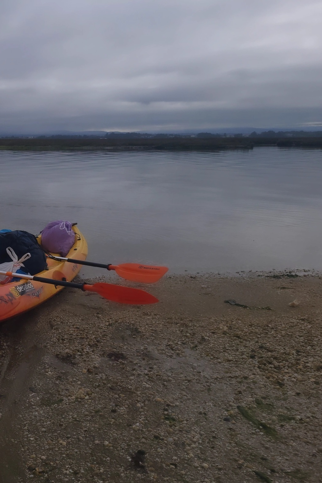 Two kayaks with orange paddles are resting on a pebbled shore by a calm, cloudy body of water.