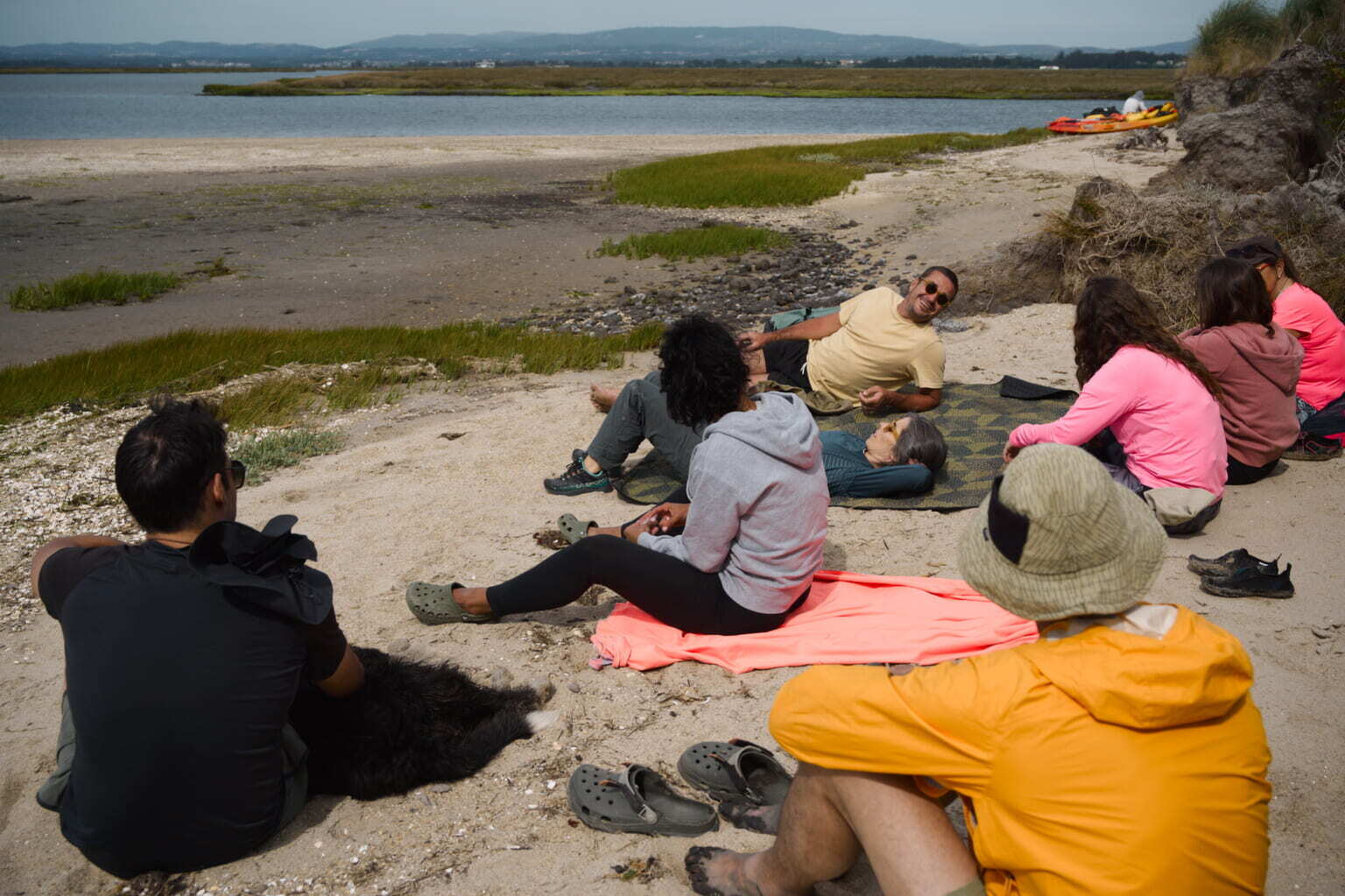 A group of people is sitting and relaxing on a sandy beach near a body of water, with some shoes and a kayak visible in the background.