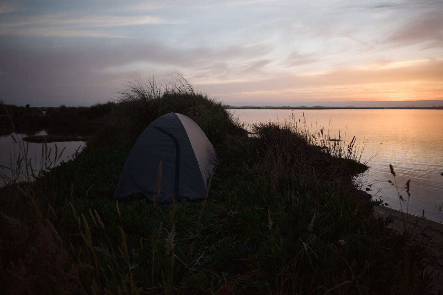 A tent is set up on a grassy riverbank at dusk, with calm water reflecting the fading light of the sky.