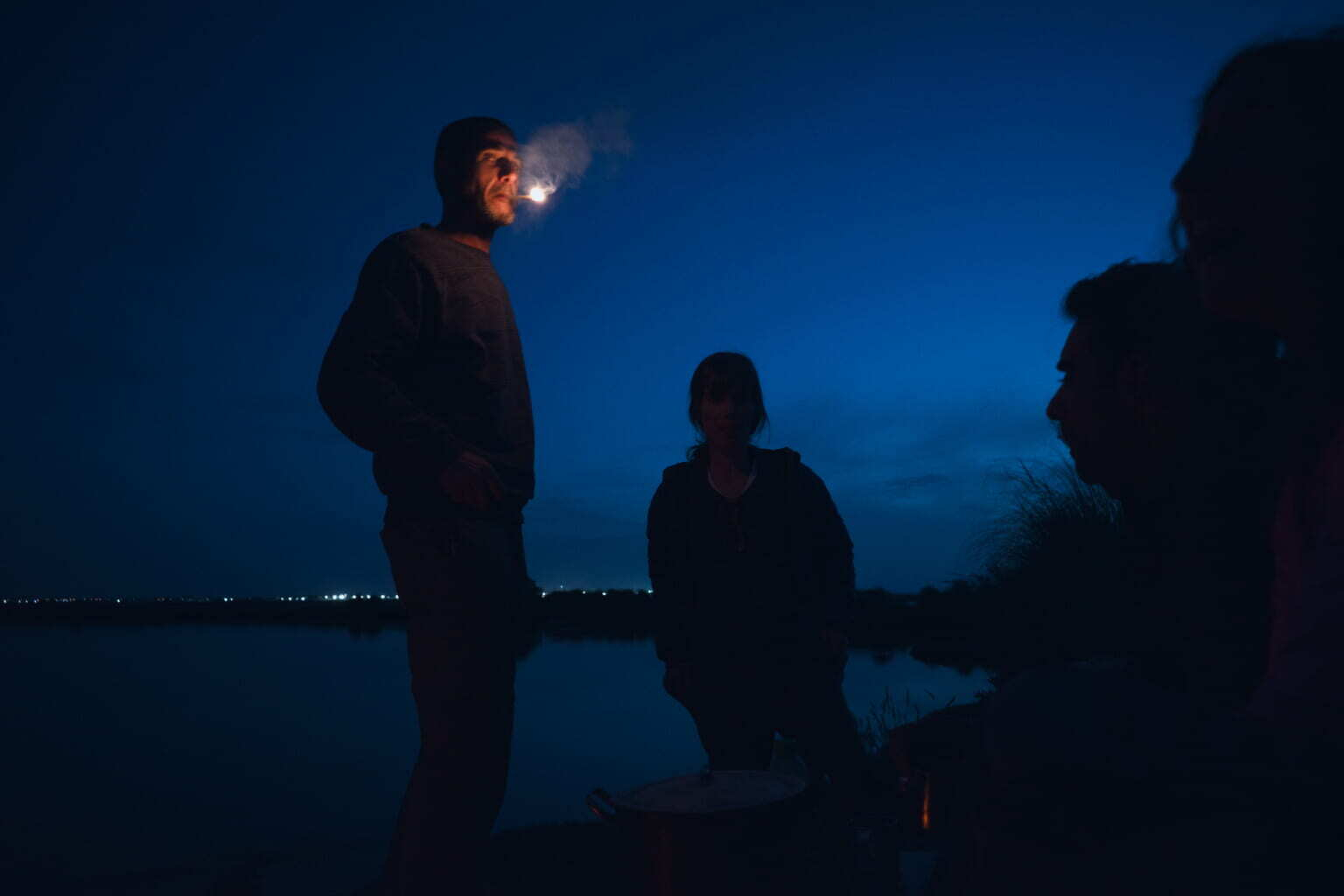 A group of people are gathered outdoors at night by the water, with one person exhaling smoke that is illuminated in the dark.