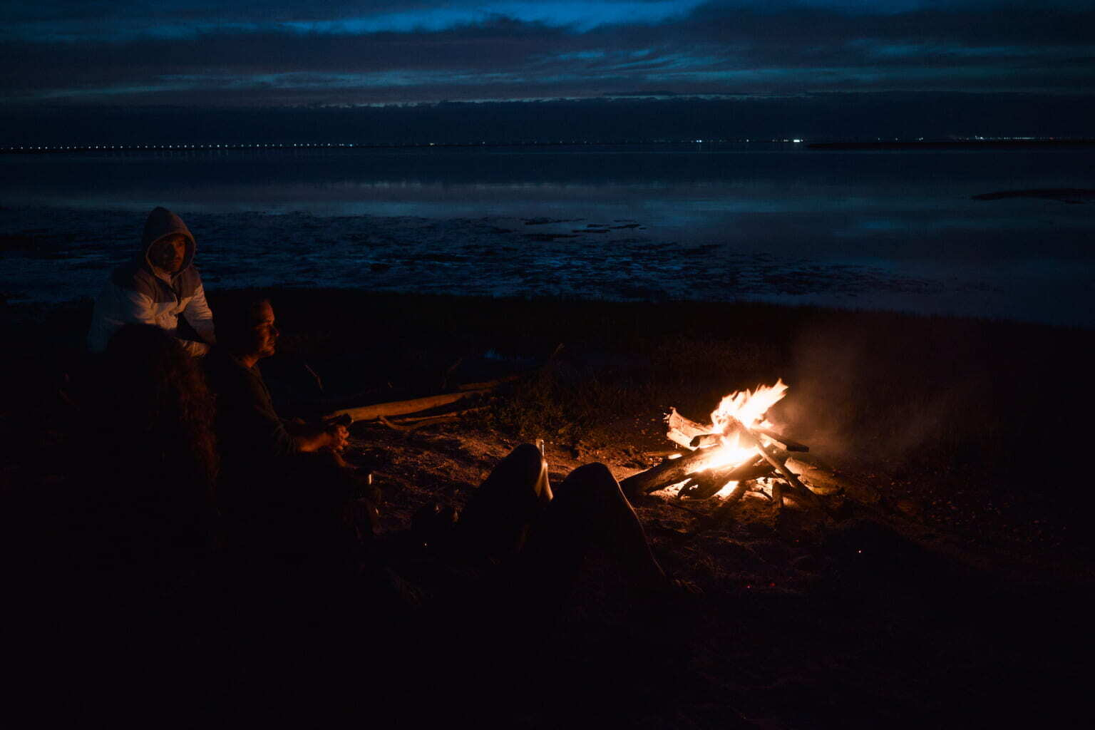 Two people are sitting by a campfire on a dark beach near the water.