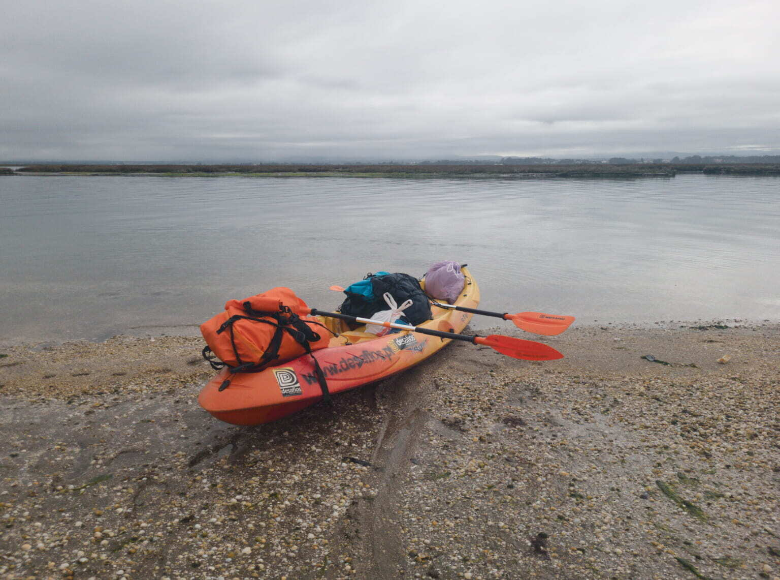 A kayak loaded with gear and two paddles rests on a pebbled shore beside calm water under a cloudy sky.