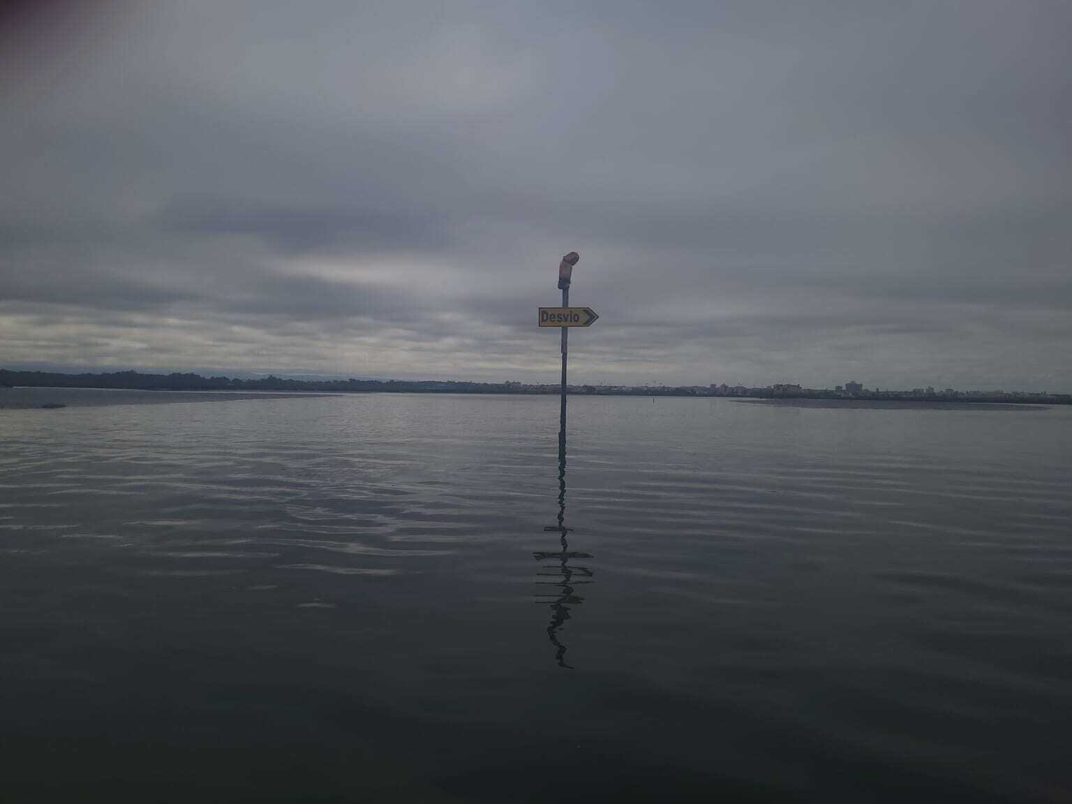 A signpost labeled Desvio stands partially submerged in calm water under a cloudy sky.