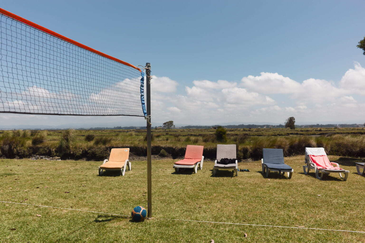 A volleyball net is set up on a grassy field with a ball at the base of the net and five lounge chairs lined up in the background.