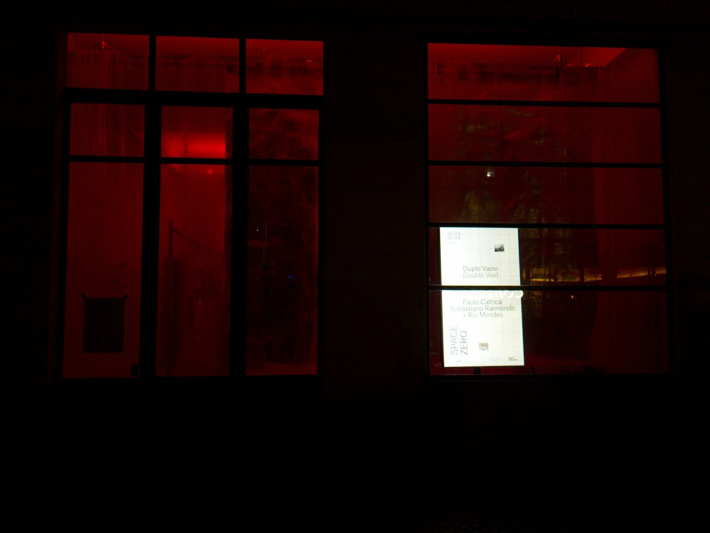 A dimly lit building exterior is illuminated by red light from inside, with a window displaying a bright digital screen showing text.