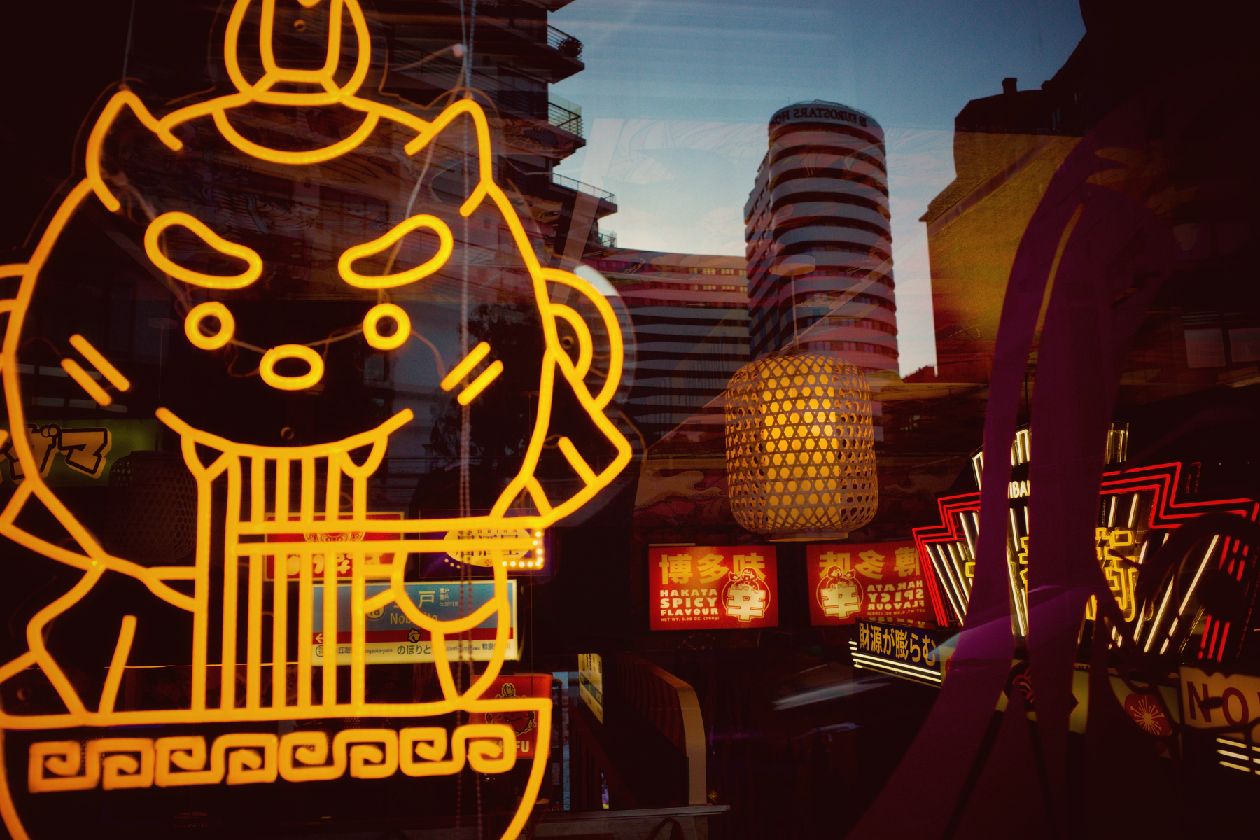 A cityscape at dusk features neon signs, including a cartoon character slurping noodles, with modern buildings in the background.