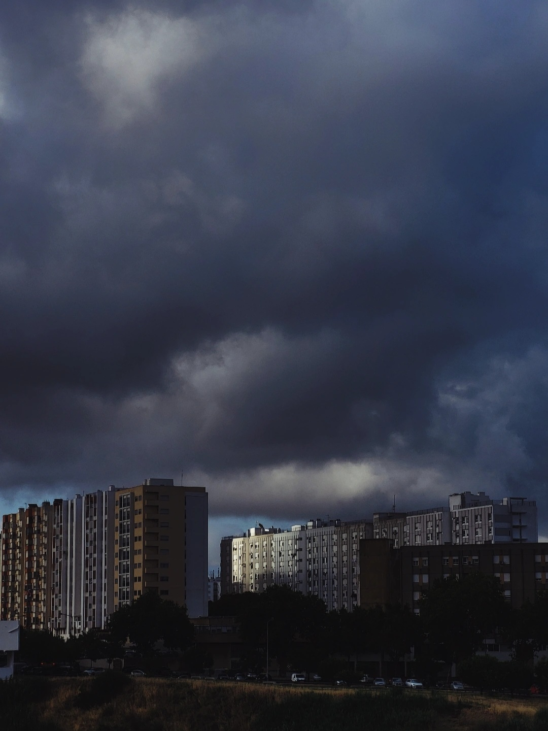 Dark, heavy clouds loom over a row of tall residential buildings, casting shadows on the cityscape below.