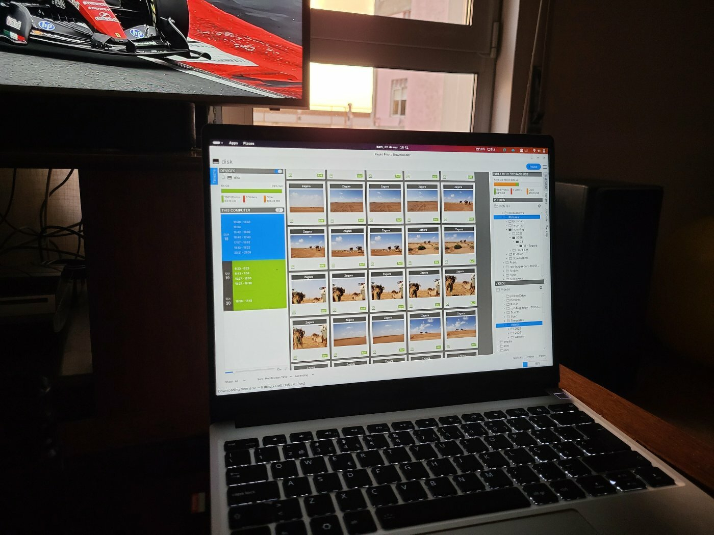 A laptop displaying a photo editing or organization application is positioned in front of a TV showing a racing event.