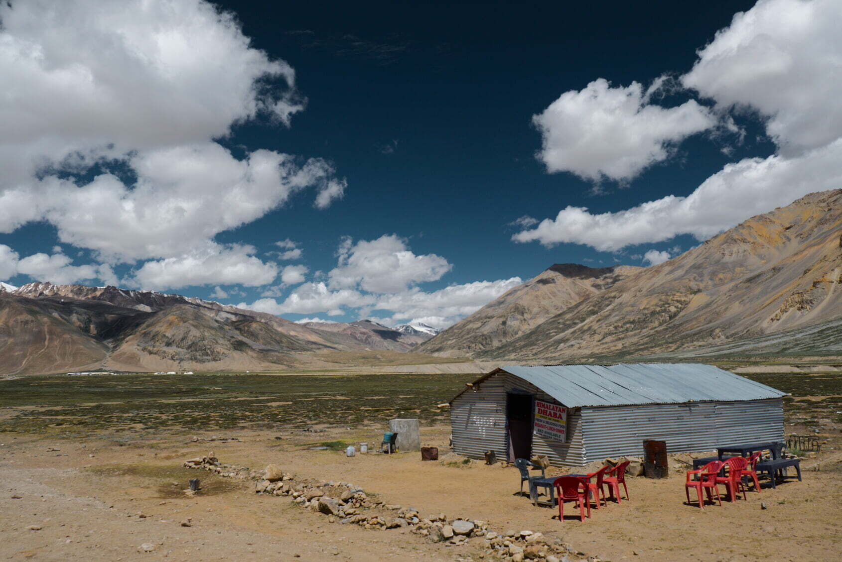 A small, rustic roadside shack with red plastic chairs is set against a vast, mountainous landscape under a partly cloudy sky.