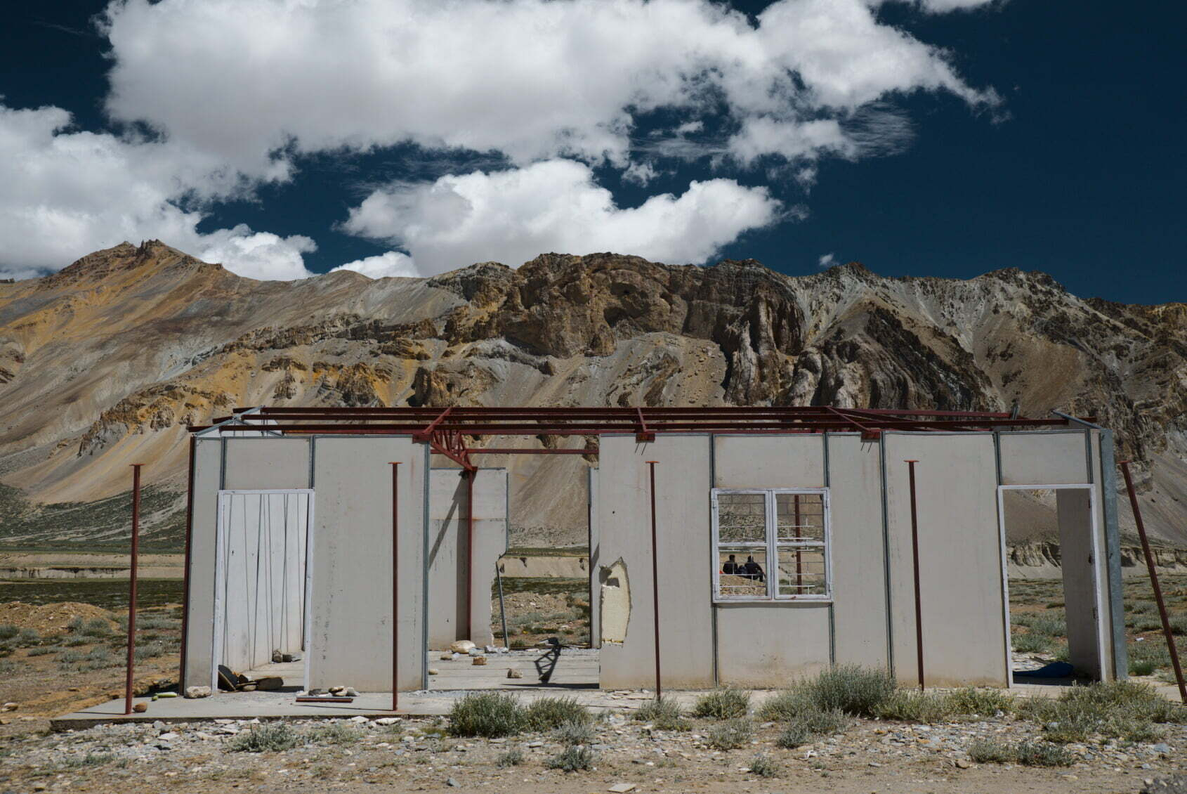 A partially constructed or abandoned building stands in a barren landscape with rugged mountains and a dramatic cloudy sky in the background.