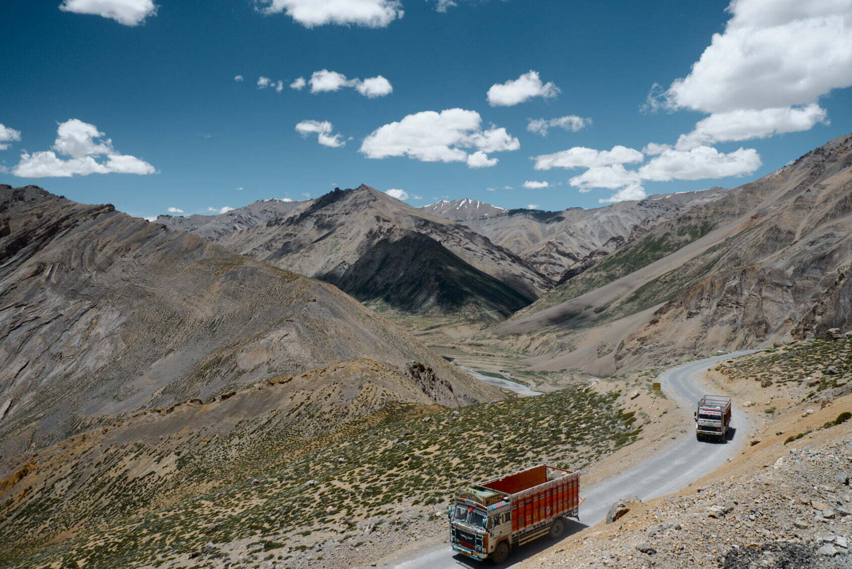 A winding mountain road with two trucks driving through a rugged, rocky landscape under a bright blue sky with scattered clouds.