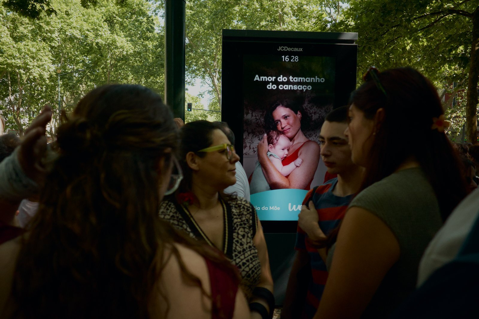 A group of people are standing near a digital display showing an advertisement with a mother holding a baby.