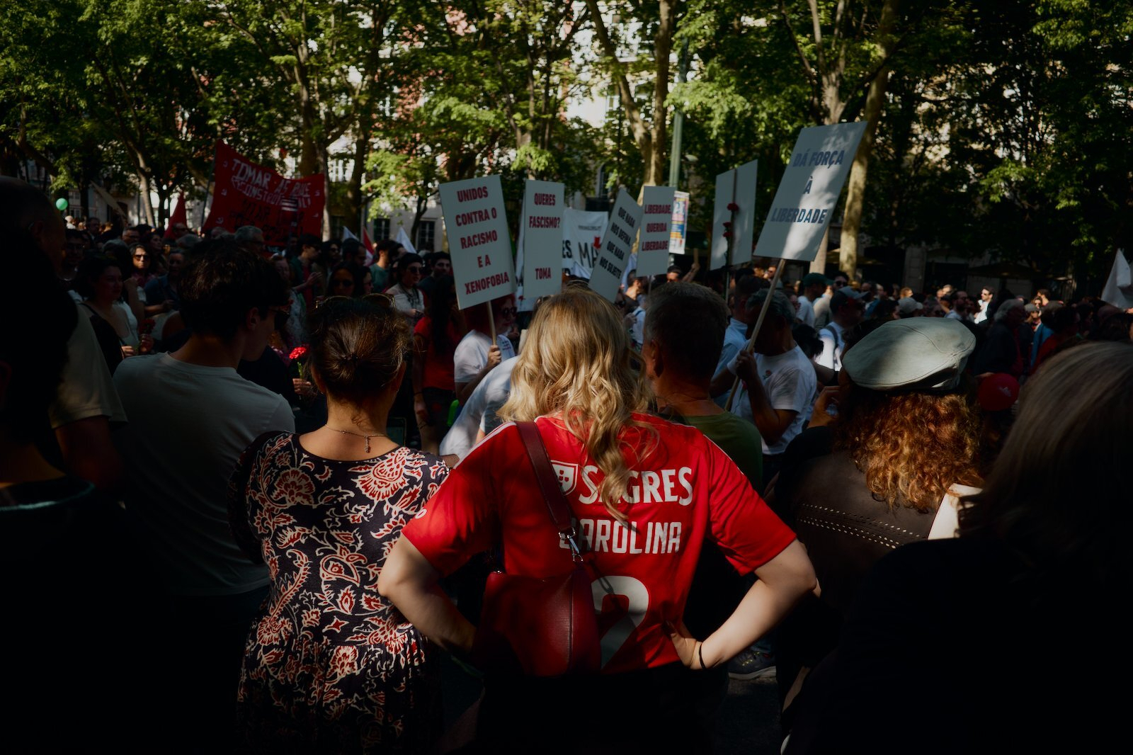 A crowd of people, some holding signs, gathers in a shaded outdoor area, with one person wearing a red sports jersey prominently in the foreground.